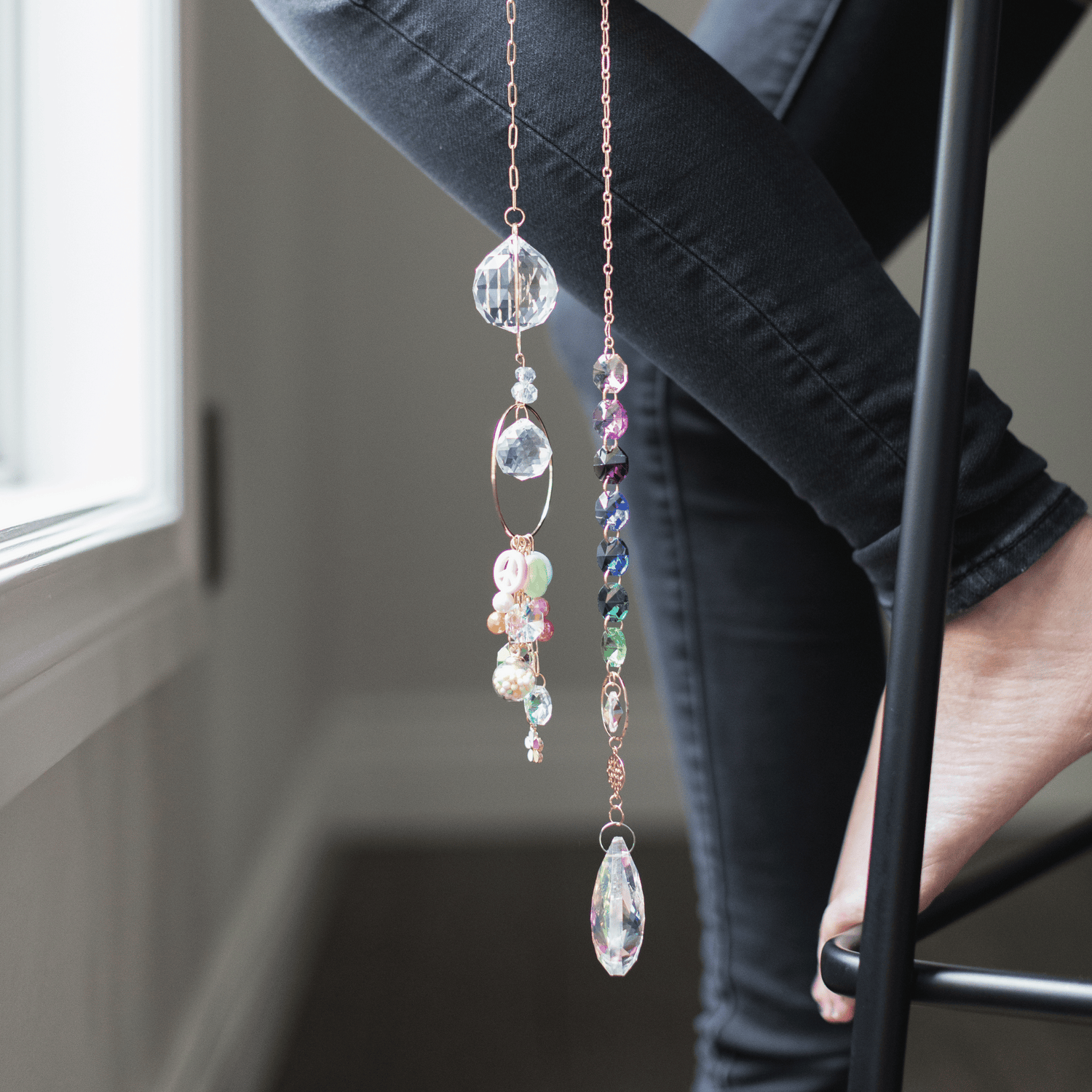 A human perched on a stool wearing denim and a bare foot. Two Prism Suncatchers are dangling in front